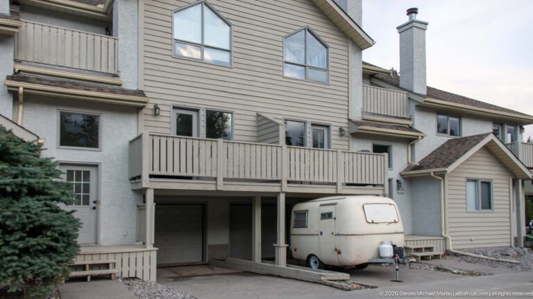 A compact travel trailer is parked beneath the balcony of a pale-sided residential building.