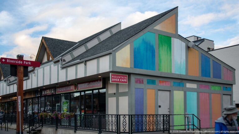 A colorful storefront with vertical turquoise, green, and orange panels stands beside a Riverside Park sign.