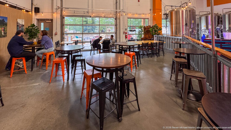 Taproom tables and stools face seated patrons, with brewing tanks visible along the right wall.