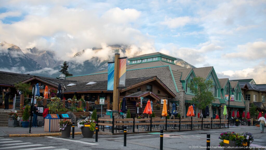 A mountain-town dining terrace with colorful umbrellas and shops sits below cloud-brushed peaks.