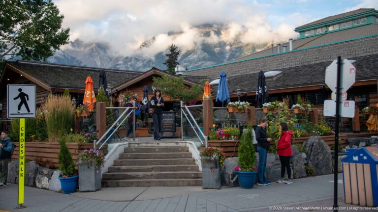 Stone steps lead to a flower-filled outdoor dining terrace with umbrellas and visitors beside a town sidewalk.