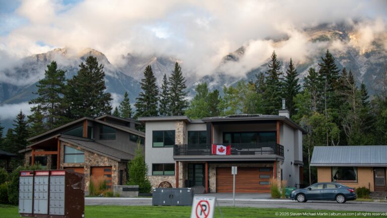 A Canadian flag hangs from a modern home’s balcony, with cloud-wrapped Rocky Mountain peaks behind it.