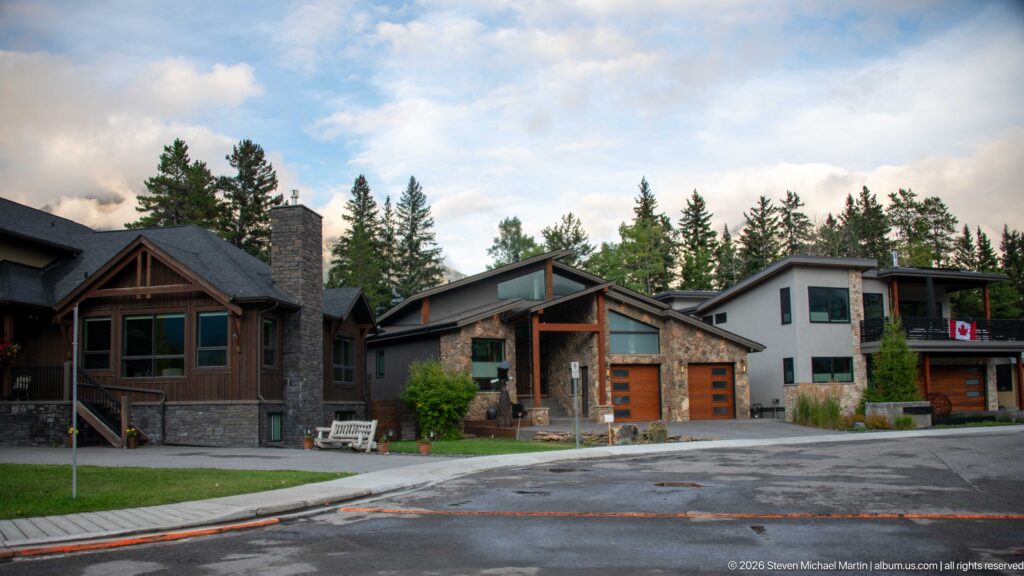 Contemporary detached homes with timber gables and stone façades line a residential street beneath a wooded mountain ridge.