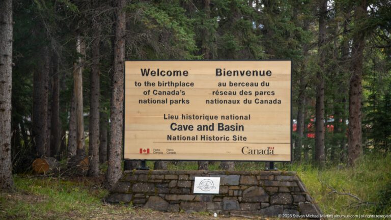 Bilingual timber welcome sign on a stone base among conifers, naming Cave and Basin National Historic Site.