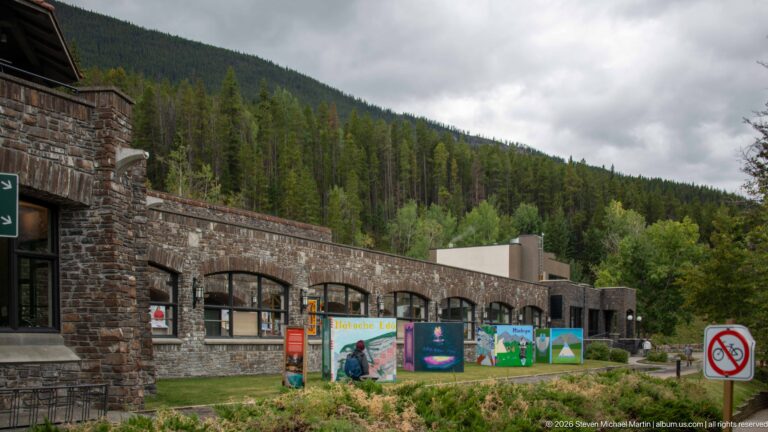 A stone arcade and colorful exhibit panels line a path beside a no-bicycles sign.