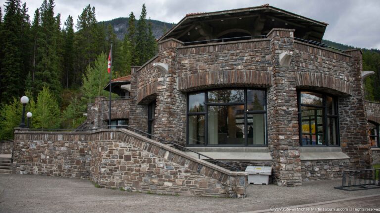 A stone visitor center with an upper balcony and Canadian flag stands among conifers.
