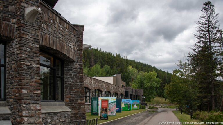 Colorful interpretive panels line the path beside a stone visitor-center façade and forested slope.