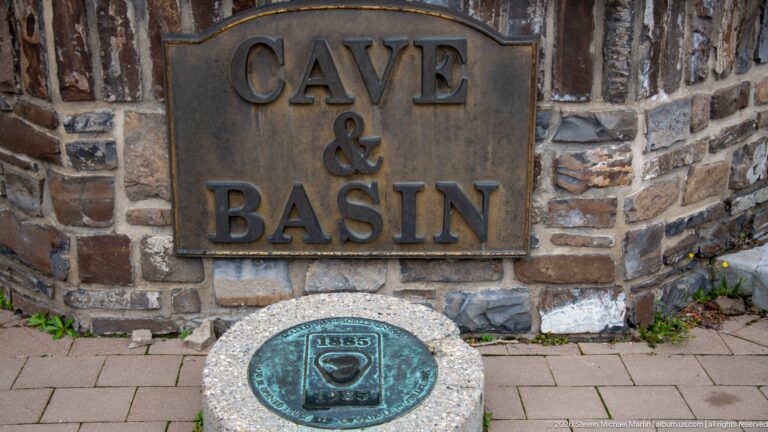 A Cave & Basin plaque hangs above an 1885–1985 medallion set into the pavement.