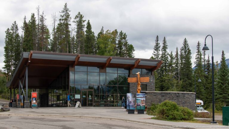 Contemporary visitor center with a broad roof, glass façade, stone walls, and a paved forecourt.
