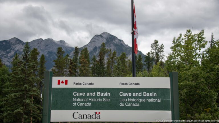 Parks Canada site sign and Canadian flag beneath rocky mountain peaks and forest.