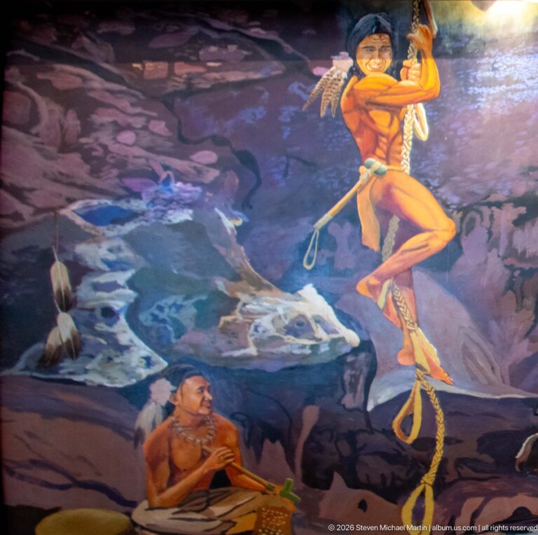 A painted Indigenous figure holds a child above a seated person amid a stylized cave landscape.