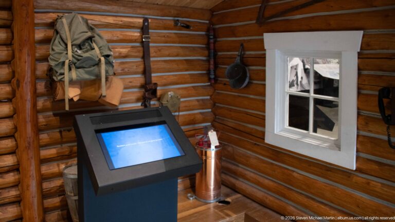 A log cabin exhibit contains hanging clothing, equipment, a touchscreen, and a small-paned window.