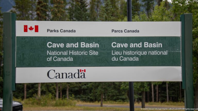 Close view of the green bilingual Cave and Basin National Historic Site sign against trees.
