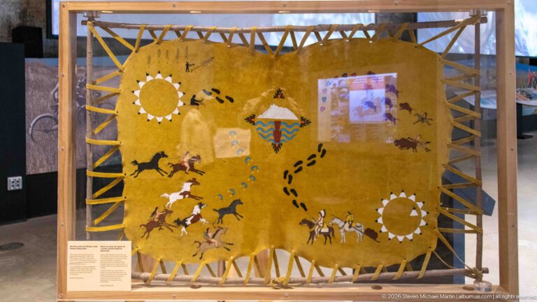 An ochre-colored hide display bears painted figures, animals, tracks, and circular motifs.