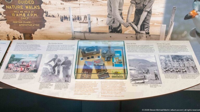 A glass case of printed memorabilia sits within a bilingual exhibit of historic visitor photographs.