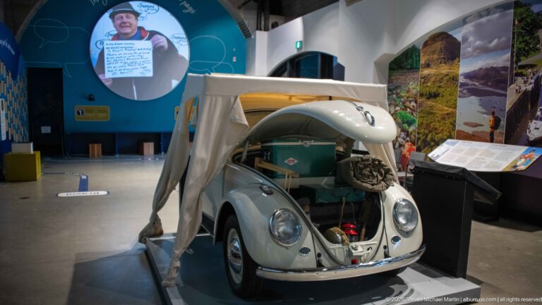 A pale green vintage Volkswagen Beetle stands in a gallery among interpretive displays.