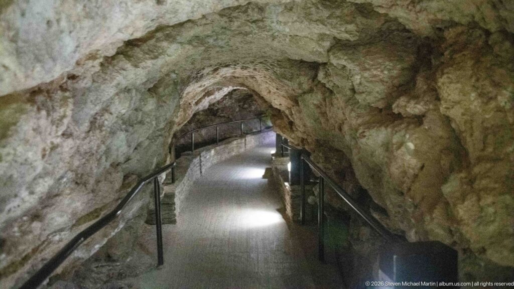 A lit boardwalk with black handrails passes through a low limestone cave passage.