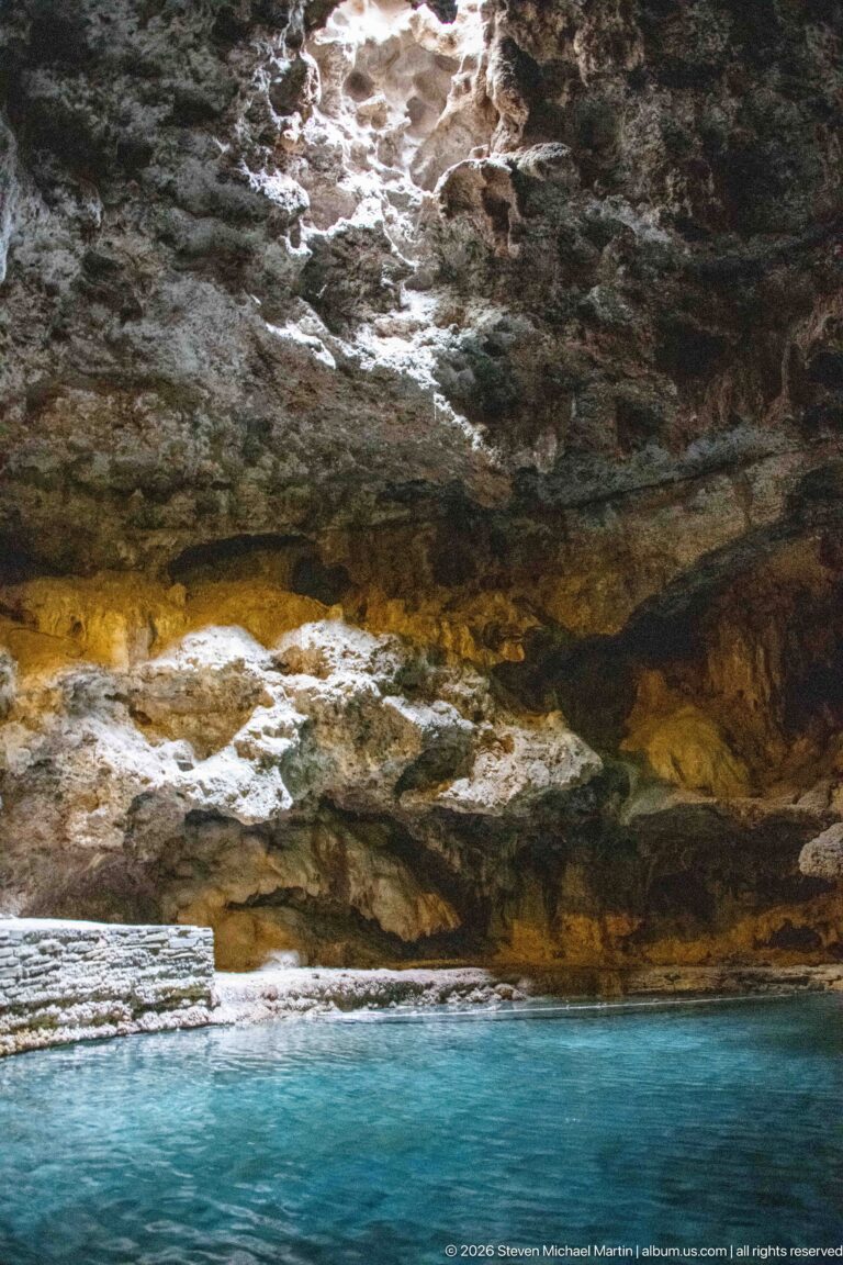 A pale mineral-stained cave wall meets the turquoise surface of the thermal pool.
