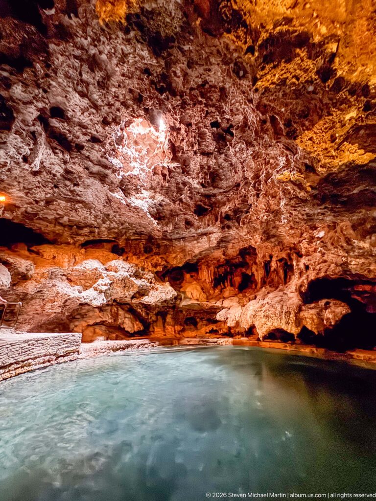 Rough pale and orange-stained cave rock extends above a narrow strip of blue-green water.
