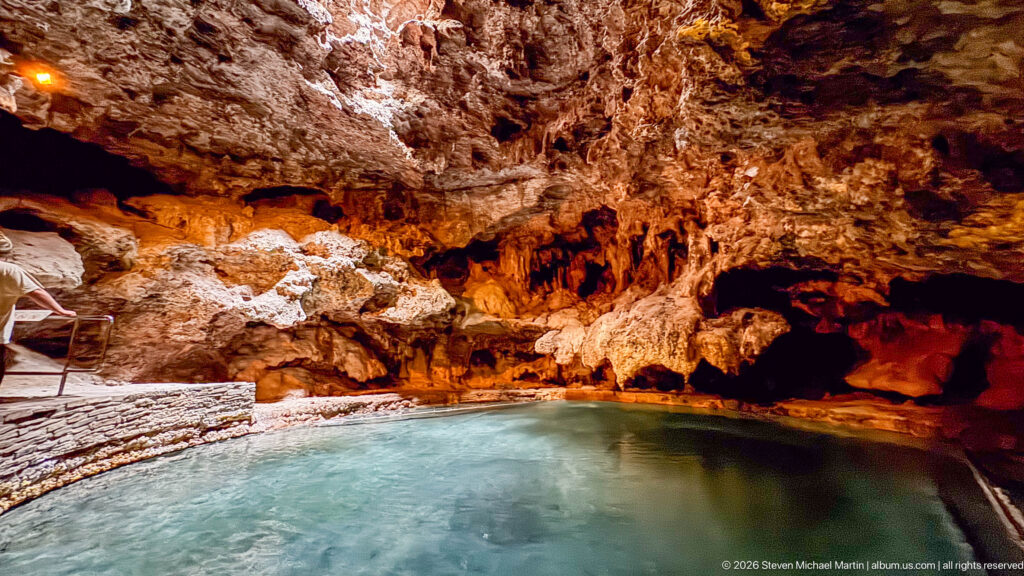 A broad turquoise cave pool lies beneath a rough illuminated ceiling and beside a stone platform.