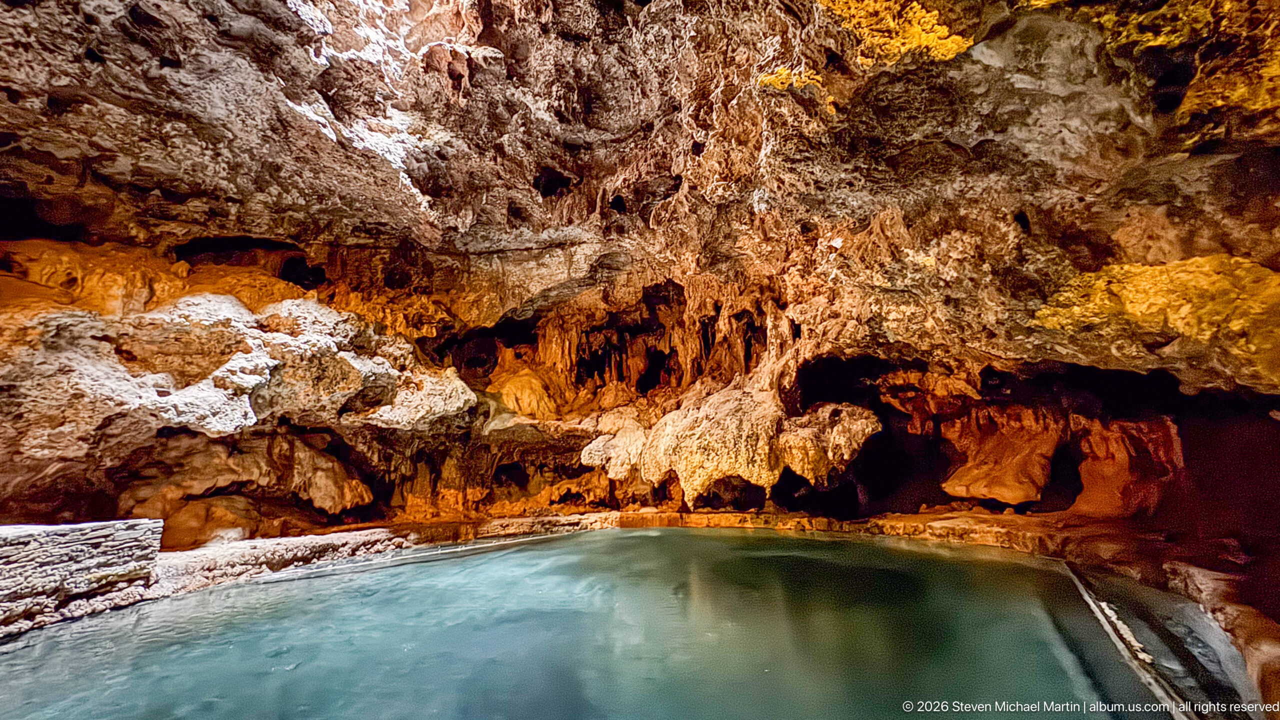 Amber-lit limestone formations overhang a turquoise pool beside a low stone walkway.