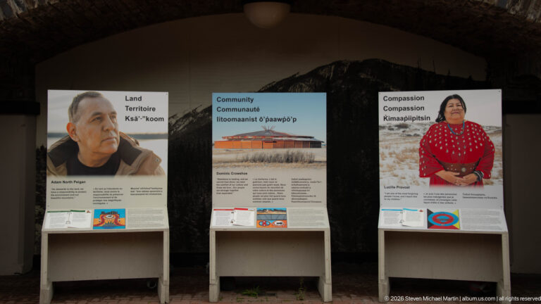 Three bilingual exhibit panels titled Land, Community, and Compassion stand against a stone wall.