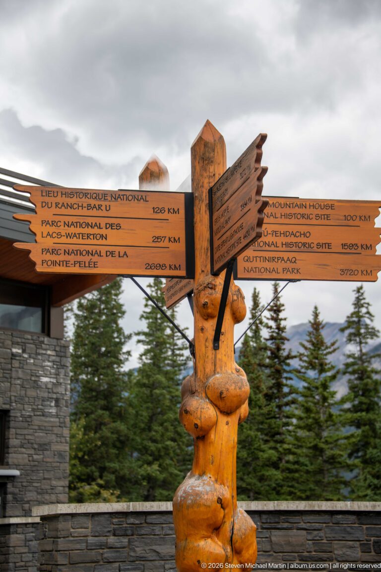 Carved wooden direction post with multiple destination arrows beside a stone wall and evergreen trees.