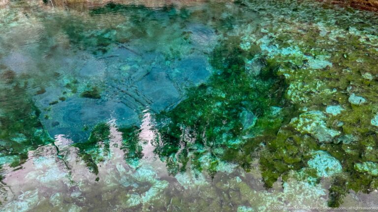 Blue-green spring water ripples over pale mineral deposits and dark patches on submerged rock.