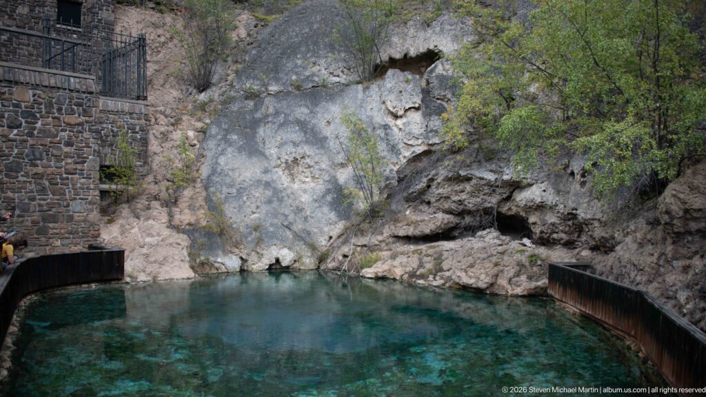 A turquoise thermal pool rests beneath a low limestone cave opening and beside a stone wall.