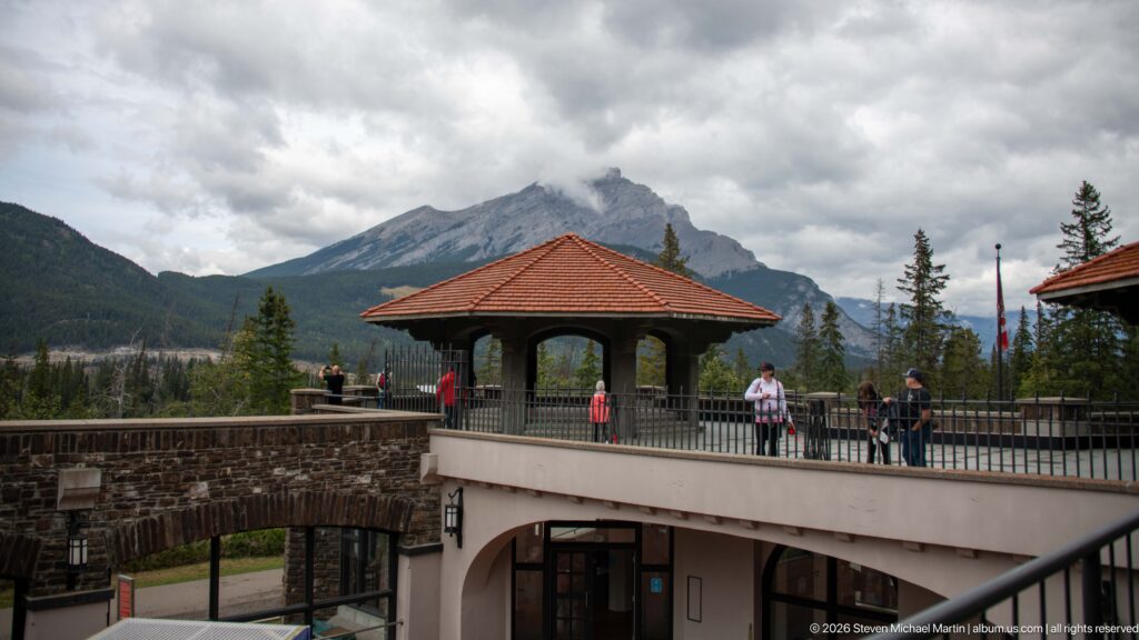 A red-roofed pavilion and stone arcade frame visitors with Rocky Mountain peaks beyond.