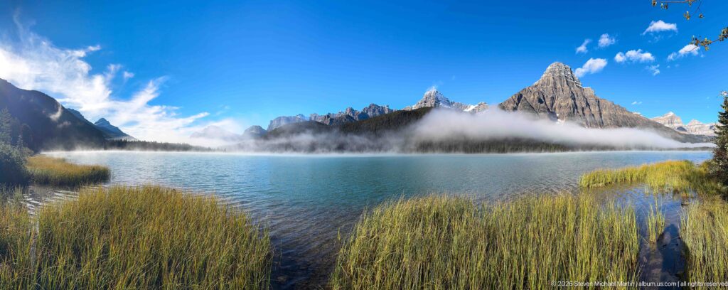 Reeds frame a lake beneath a pointed summit and cloud-draped forested shore, with a small visitor at right.