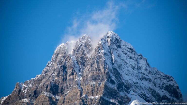 A sharp snow-covered summit rises into drifting cloud above steep exposed rock.