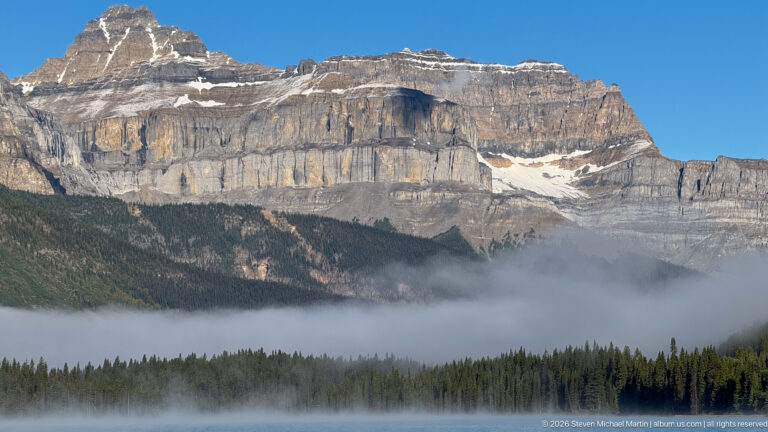 A broad layered escarpment rises above conifers and a lake, with fog crossing the lower slope.