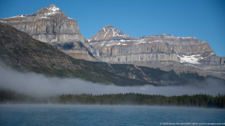 A calm lake lies beneath layered mountains as a low fog bank drifts over the conifer shore.