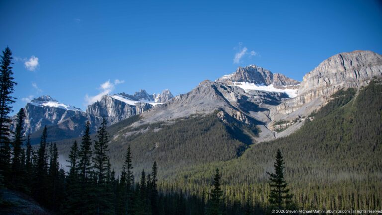 A pale layered mountain with snow patches rises above a green valley and dark conifer forest.