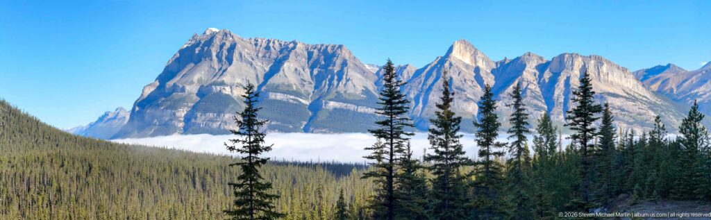 A jagged snow-dusted summit rises above dark conifers and a low cloud bank.