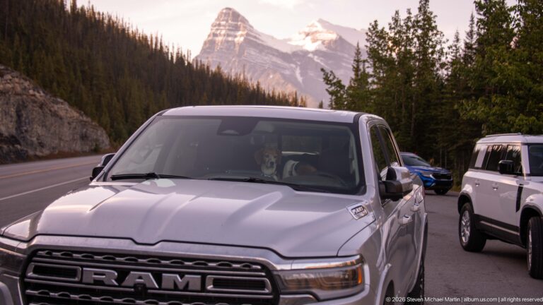 A white Ram pickup and another vehicle pause on a mountain road beneath a steep snow-streaked peak.