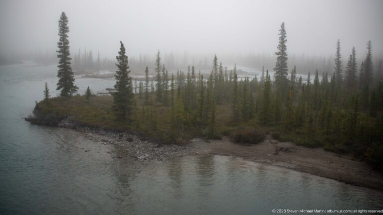 A narrow gravel island with scattered spruce divides misty turquoise river channels.