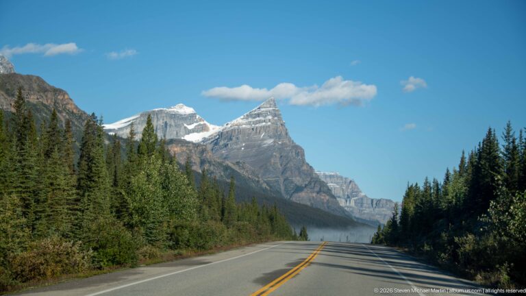 A two-lane highway runs through conifers toward distant snow-covered peaks beneath a blue sky.