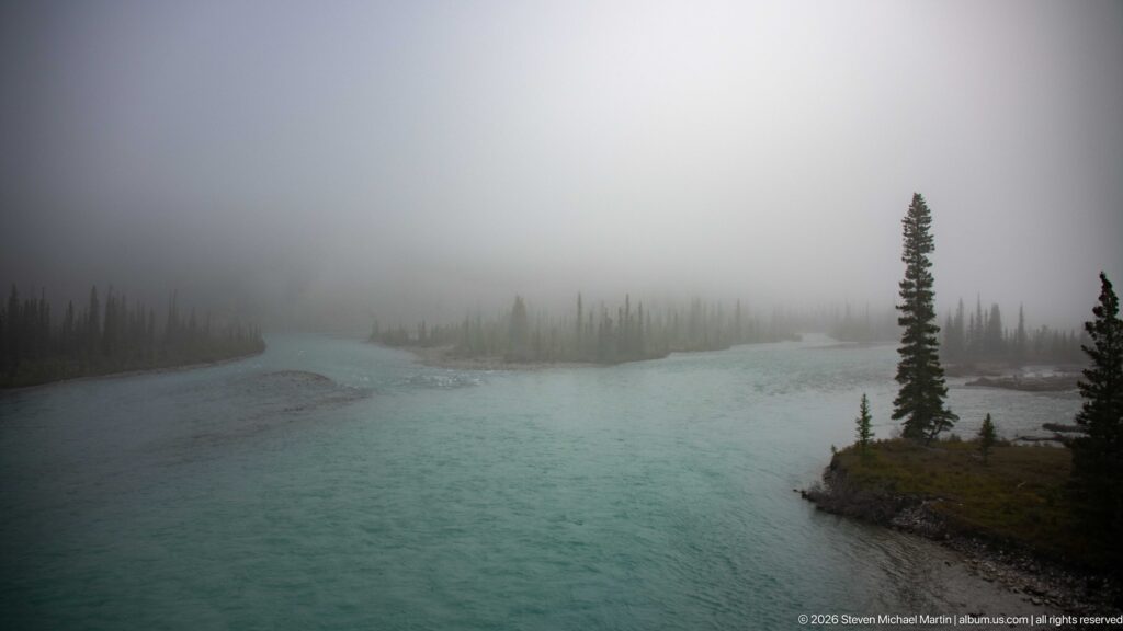 A turquoise river curls around a spruce-covered gravel point as fog obscures the far bank.