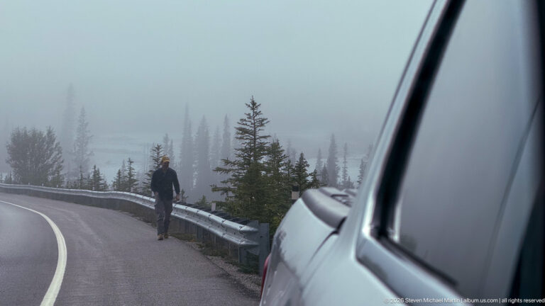 A traveler walks beside a guardrail on a foggy mountain highway, with a white vehicle in the foreground.