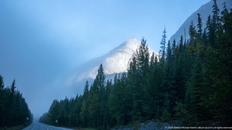 A mountain highway curves through dense conifers toward a peak partly hidden by drifting cloud.