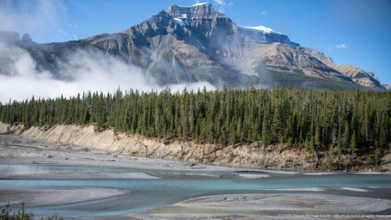 A stratified mountain partly veiled by cloud rises above forest, gravel bars, and a turquoise river.