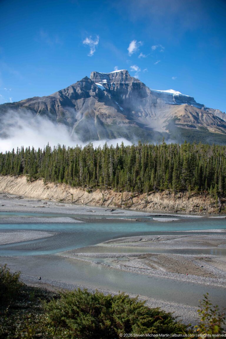 A turquoise river and gravel bars lie below a long layered mountain ridge and conifer forest.