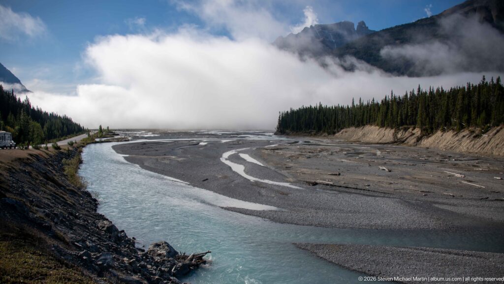 A turquoise river curves beside a broad gravel plain beneath cloud and dark conifer forest.