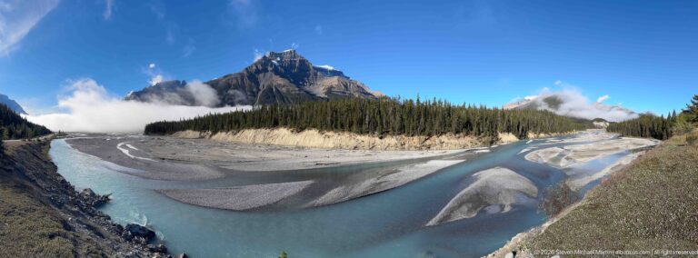 A braided turquoise river curves through gravel bars below conifer-covered slopes and a jagged peak.