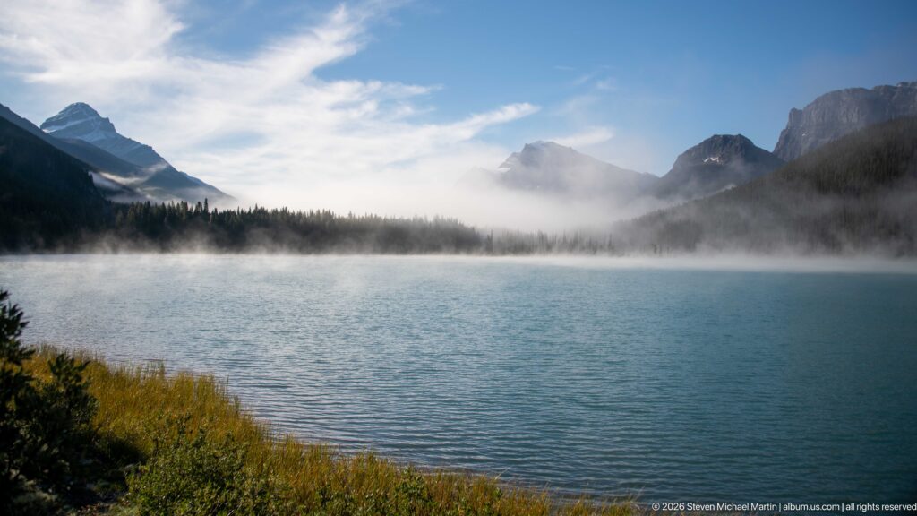 A broad lake extends toward a wooded shore and mist-softened peaks beneath broken cloud.