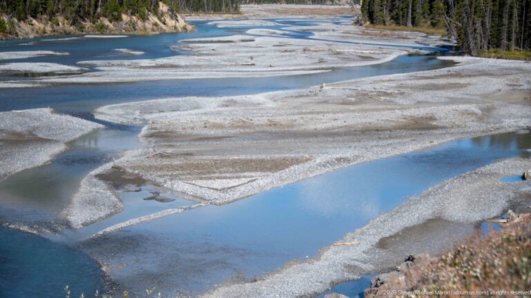 A wide braided river crosses a gravel floodplain in several turquoise channels and shallow pools.
