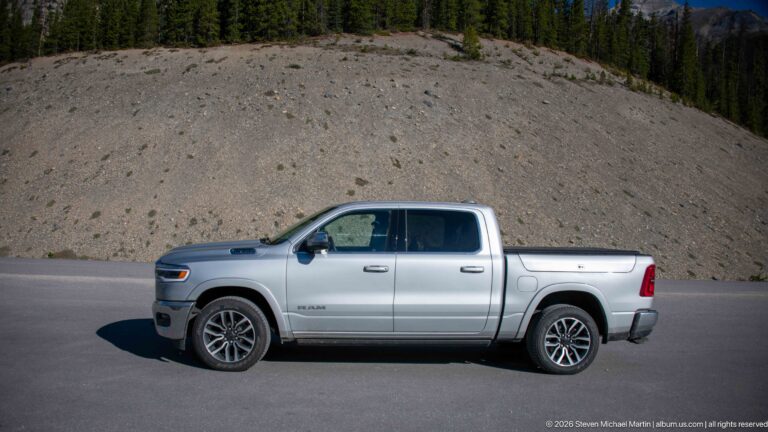 A silver four-door Ram pickup is parked on pavement below a gravel slope and conifer ridge.