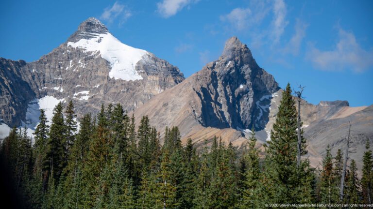 A pointed mountain with snow patches rises above dark conifers beneath blue sky and low cloud.